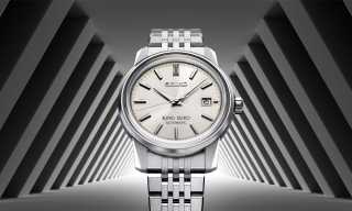 King Seiko Automatic armbandsur i rostfritt stål med silverfärgad urtavla och datumvisning, presenterad mot en symmetrisk arkitektonisk bakgrund i grå toner.