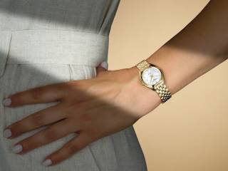 En närbild av en arm som bär en Certina DS-2 Lady-klocka helt i guld med ett länkarmband och vit urtavla. Personen bär en ljusbeige klänning med bälte.