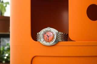 En Doxa SUB 300 klocka med en orange urtavla och ett stållänkarmband, placerad i en orange hylla.