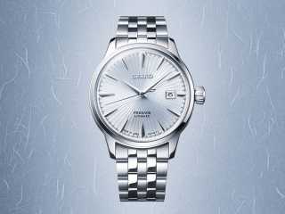 En Seiko Presage automatisk herrklocka i silver med en ljus, mönstrad urtavla och datumvisning, mot en ljusblå, texturerad bakgrund.