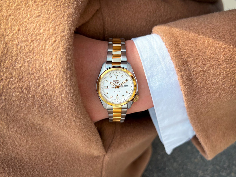 En närbild av en arm som bär en Seiko-klocka med länkarmband i silver och guld och en vit urtavla. Personen bär en vit skjorta och en brun eller beige jacka.