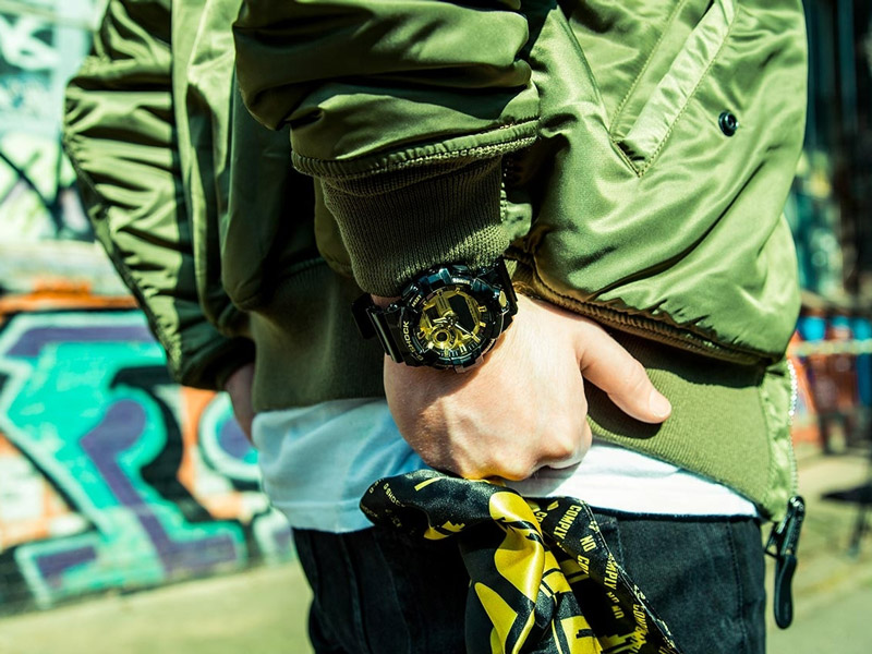 En man som står upp med en grön jacka med händerna bakom ryggen med en svart klocka från Casio med gula detaljer.