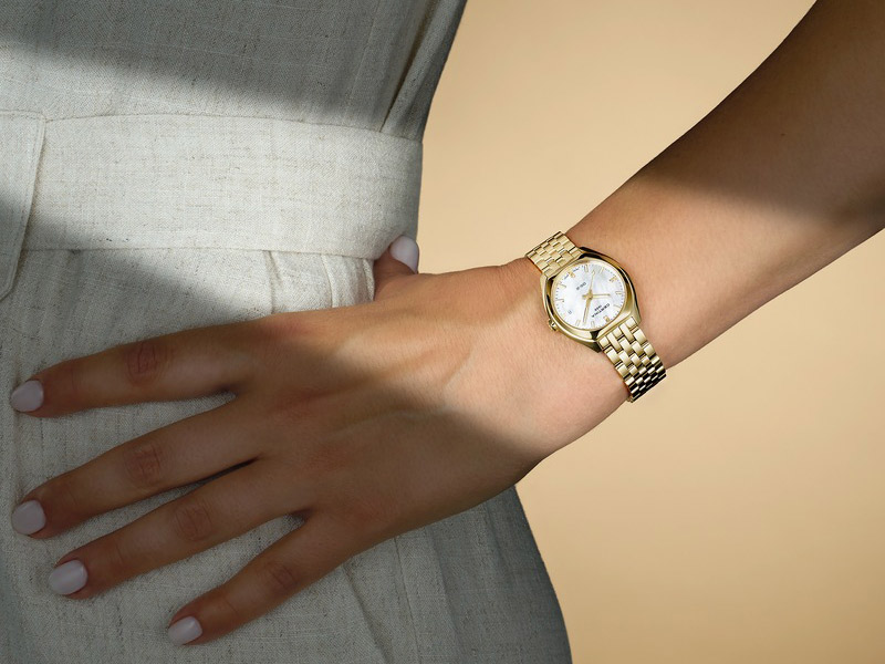 En närbild av en arm som bär en Certina DS-2 Lady-klocka helt i guld med ett länkarmband och vit urtavla. Personen bär en ljusbeige klänning med bälte.