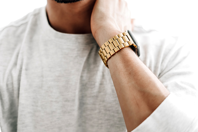 En man som håller sig för nacken och på handleden bär han en klocka med ett klockarmband i guldfärgat stål.