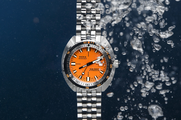 En Doxa SUB 1500 klocka med en orange urtavla och ett stållänkarmband, nedsänkt i vatten med bubblor runt omkring, mot en mörkblå bakgrund.