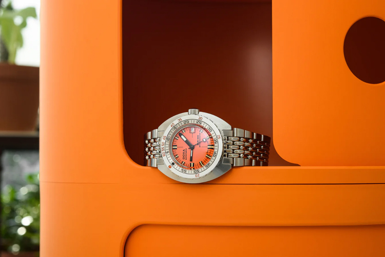 En Doxa SUB 300 klocka med en orange urtavla och ett stållänkarmband, placerad i en orange hylla.