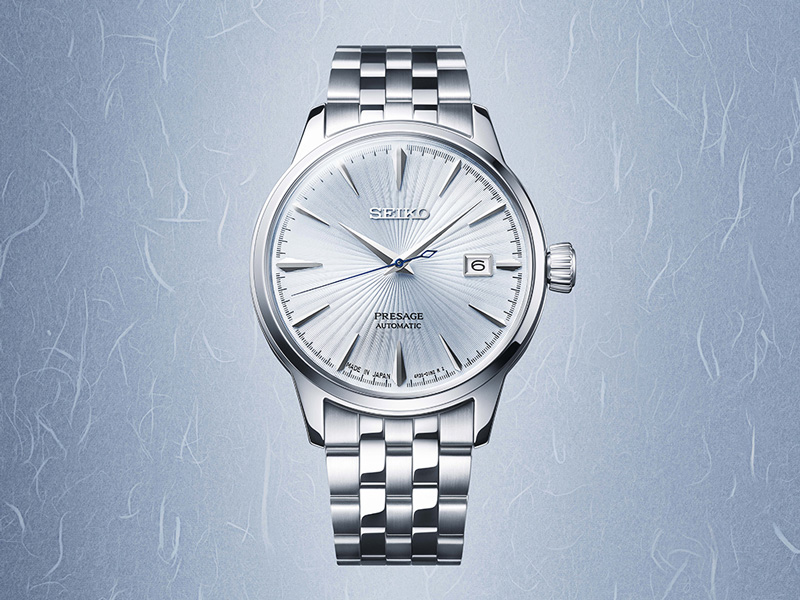 En Seiko Presage automatisk herrklocka i silver med en ljus, mönstrad urtavla och datumvisning, mot en ljusblå, texturerad bakgrund.
