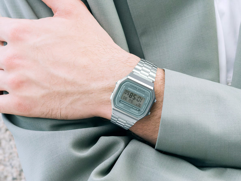 En Casio Vintage digital klocka med silverfärgat länkarmband och en grå urtavla, sitter på en handled. Personen bär en ljusgrå kavaj.