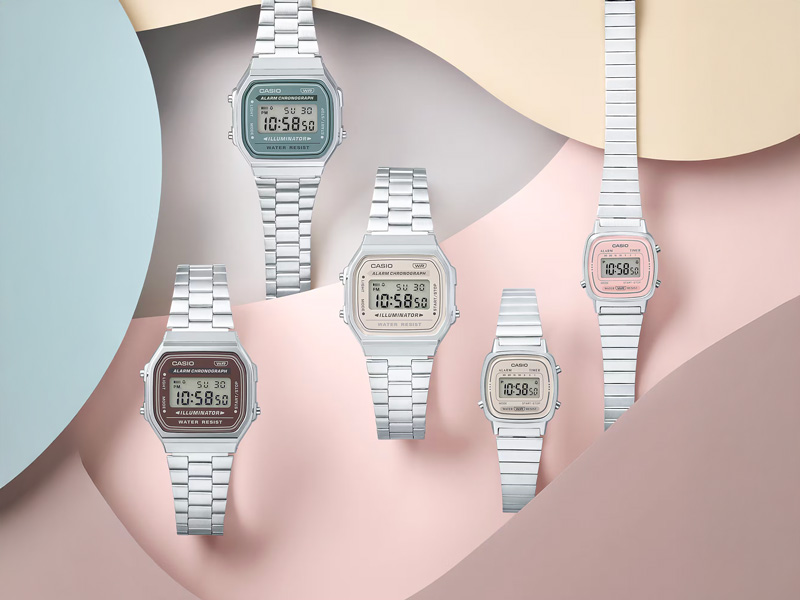 Fem Casio Vintage digitala klockor i olika färger, inklusive silver med grå urtavla, silver med ljusbeige urtavla, silver med brunröd urtavla, samt en mindre modell med silverlänk och rosa urtavla. Klockorna är arrangerade på en bakgrund med vågiga former i pastellfärger som rosa, beige och ljusblått.